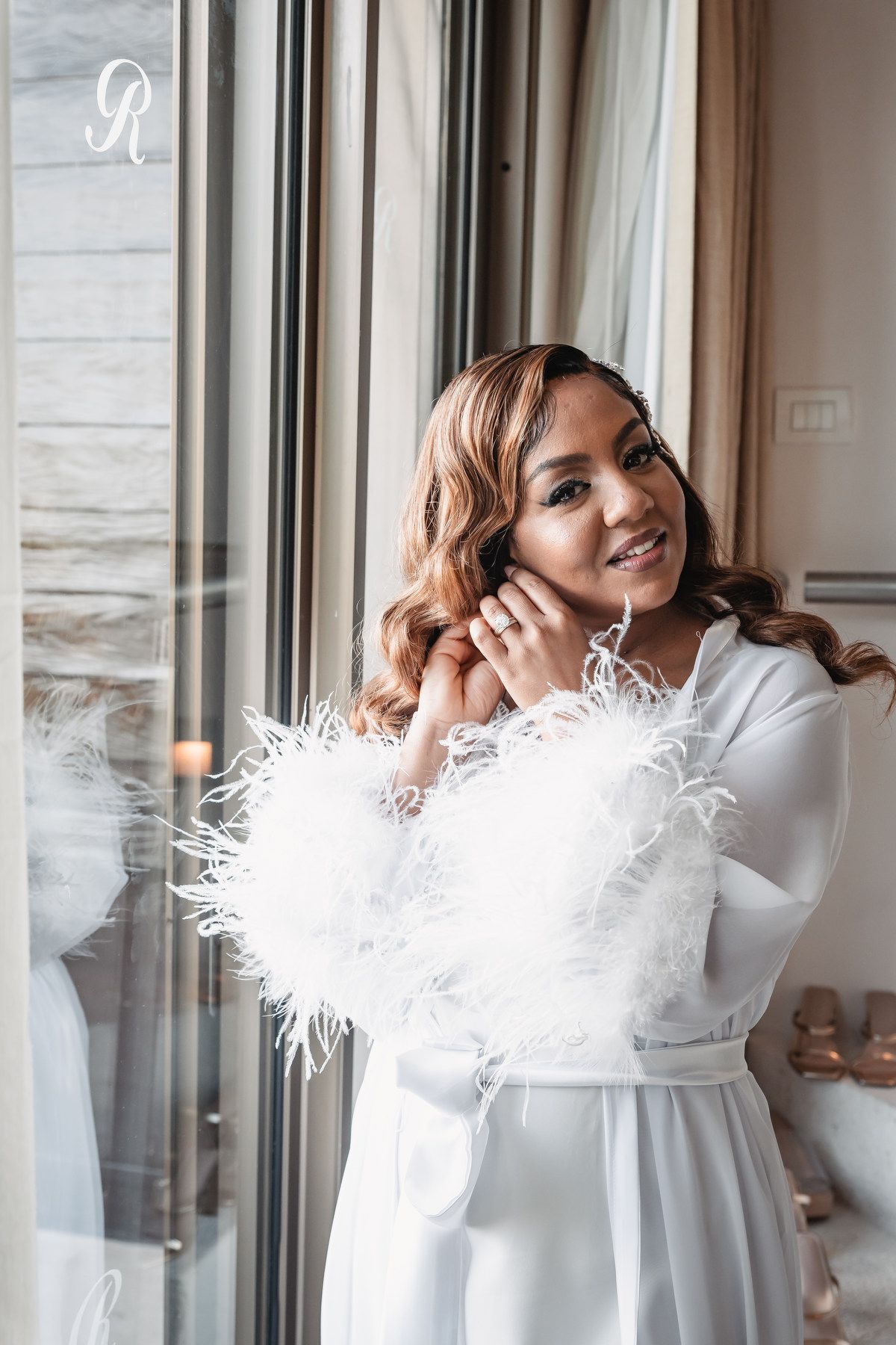 Smiling bride in white feather-trimmed robe adjusting earrings by a bright window.