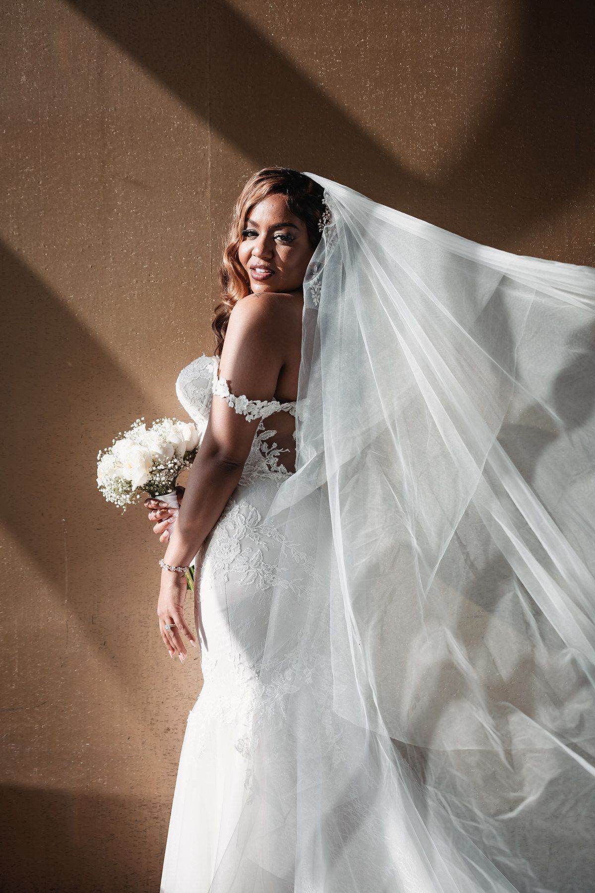 Elegant bride in lace mermaid wedding gown with flowing veil holding white bouquet against warm wall.