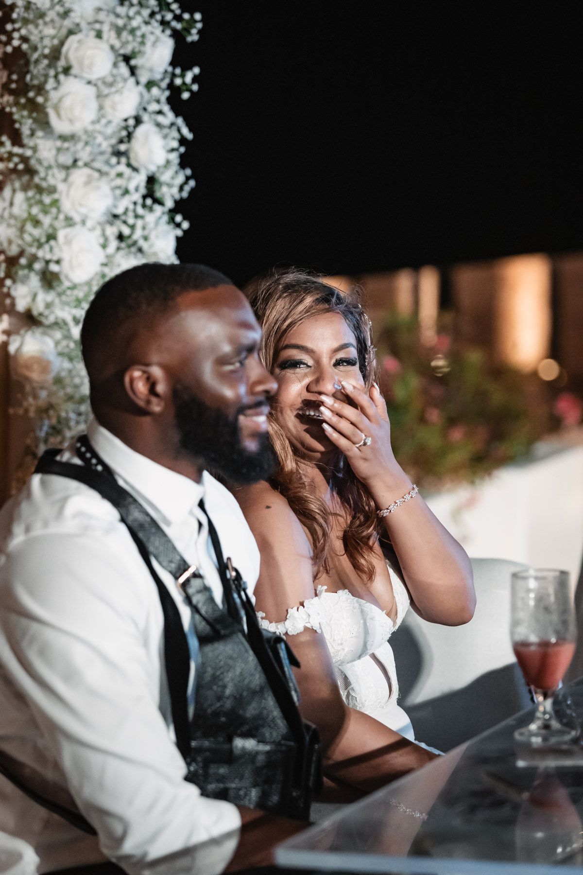 Bride and groom laugh together at their wedding reception table surrounded by floral arrangements.