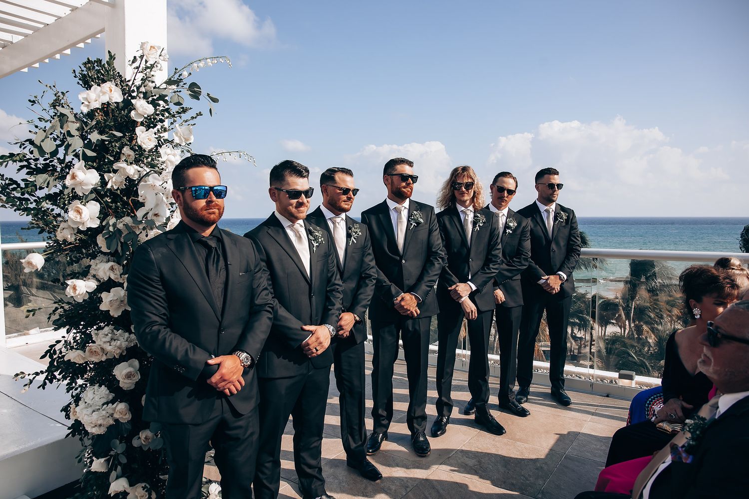 Wedding party in black suits standing in a line outdoors against a bright blue sky and ocean backdrop.