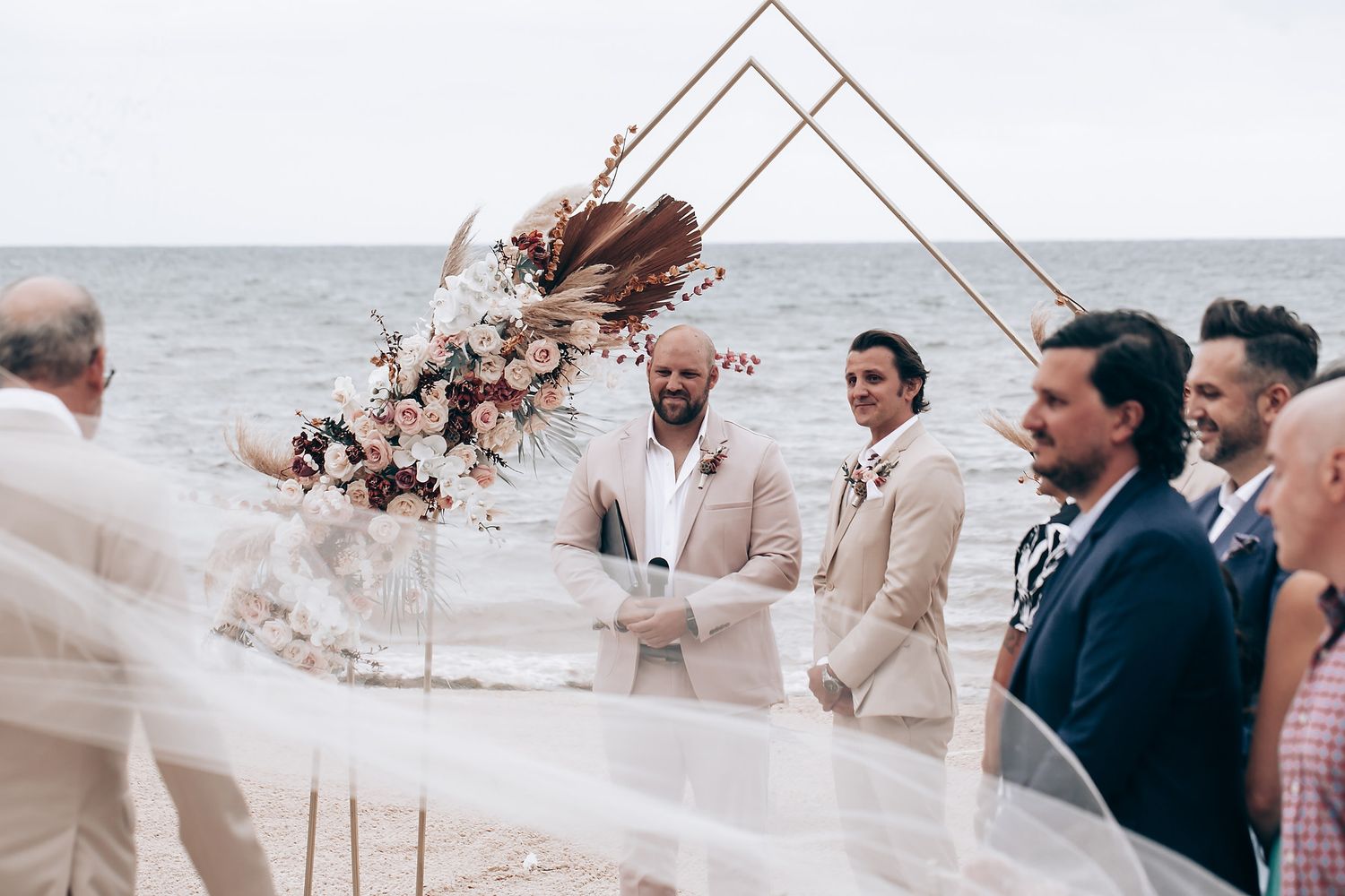 Geometric arch decorated with dried flowers frames an oceanfront wedding ceremony.