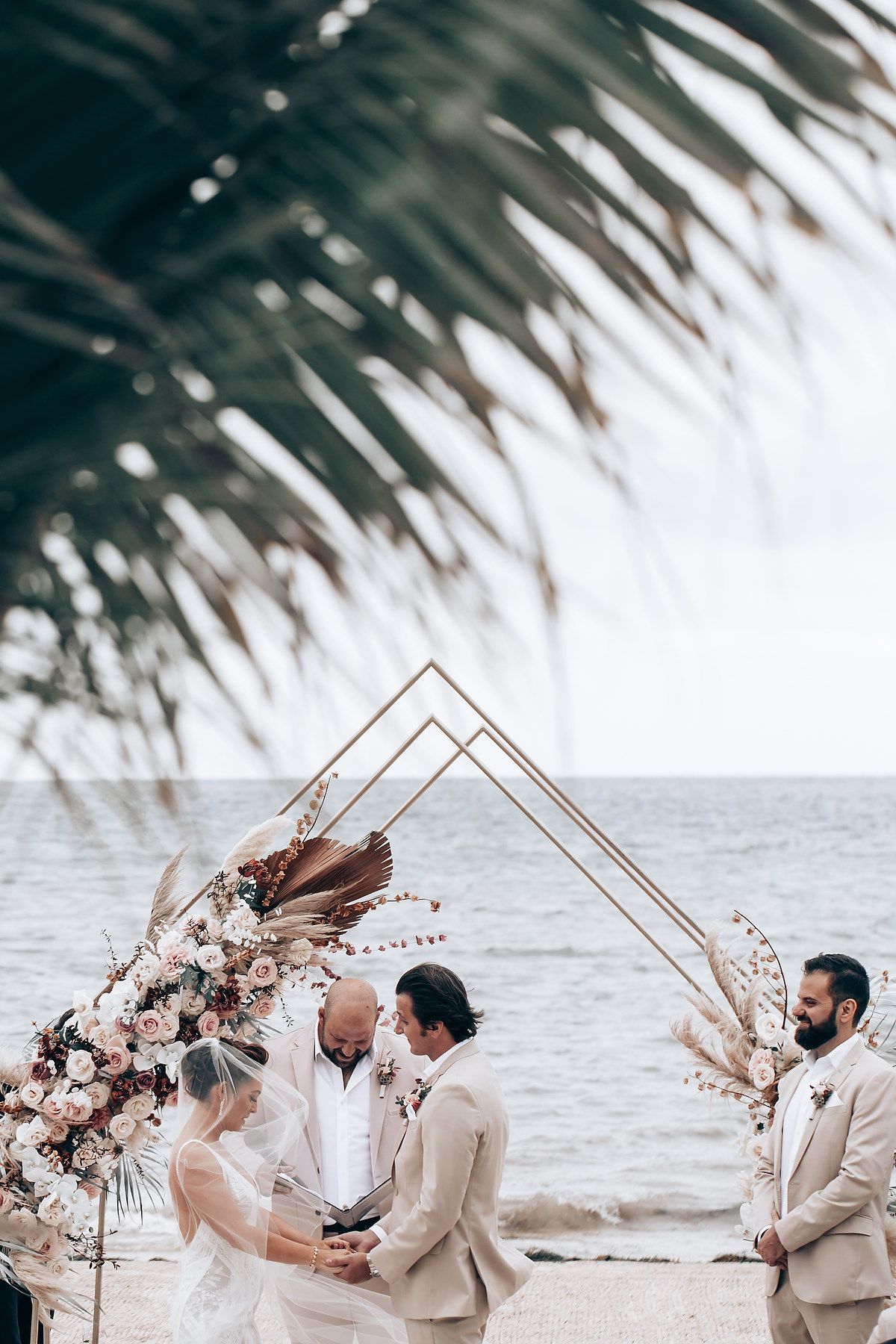 Palm fronds frame a beachside wedding ceremony with a modern geometric arch and floral arrangements.