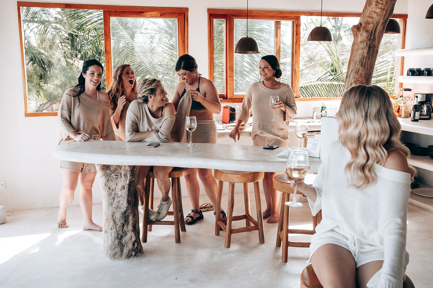 A group of people in light colored clothing gather and dance in a bright, airy indoor space with large windows.