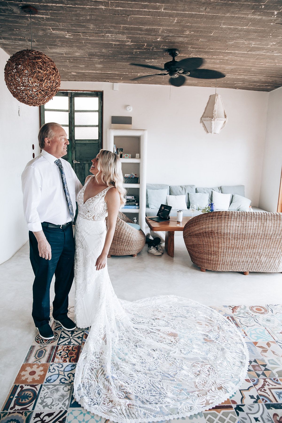 Wedding couple in white attire standing in spacious room with patterned rug and woven furniture.