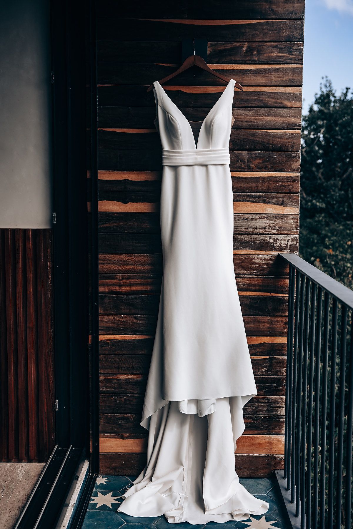An elegant white wedding dress hangs against a rustic wooden wall backdrop with natural lighting.