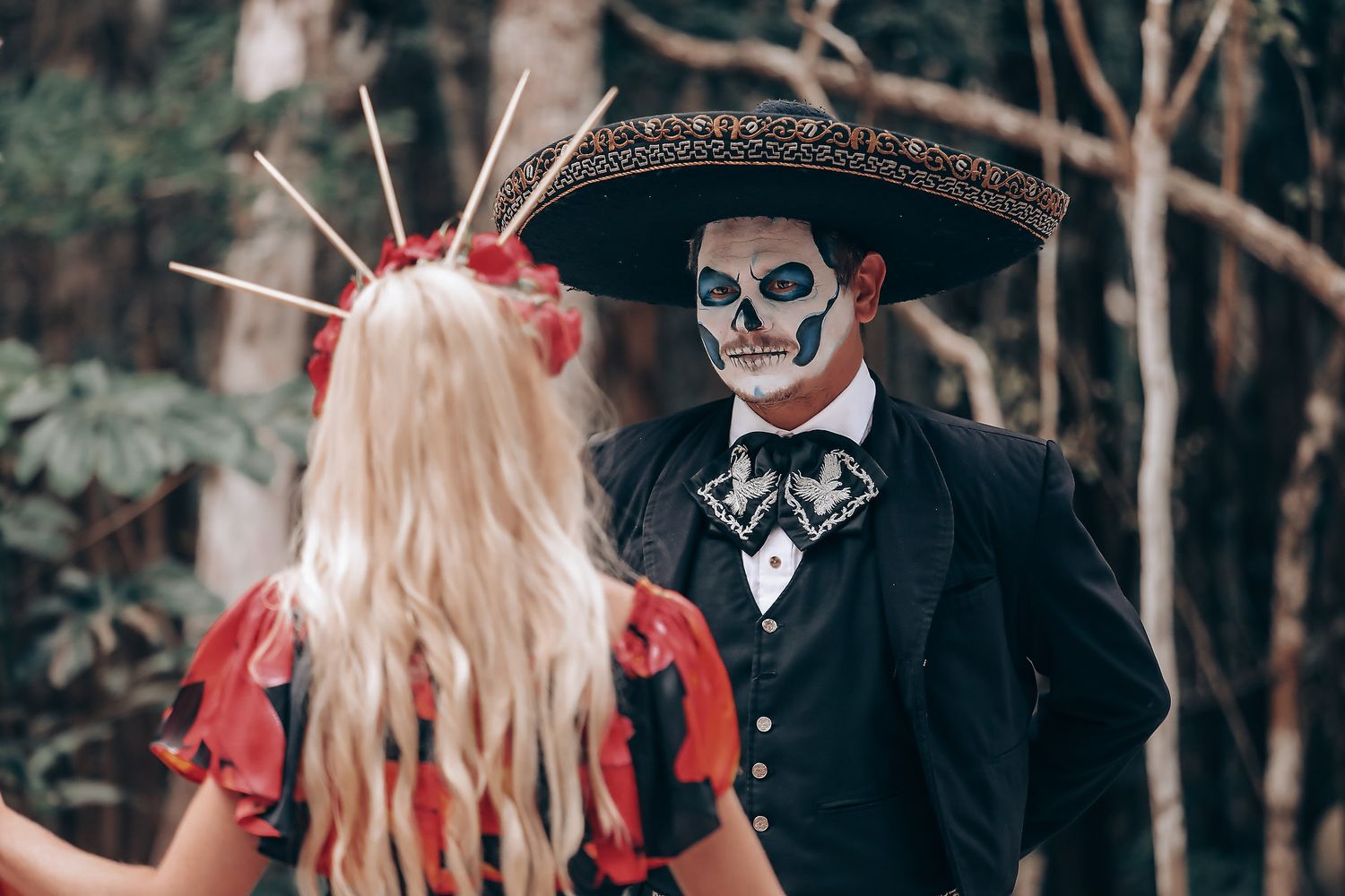 Dramatic Day of the Dead themed couple poses in black and red attire against forest backdrop.