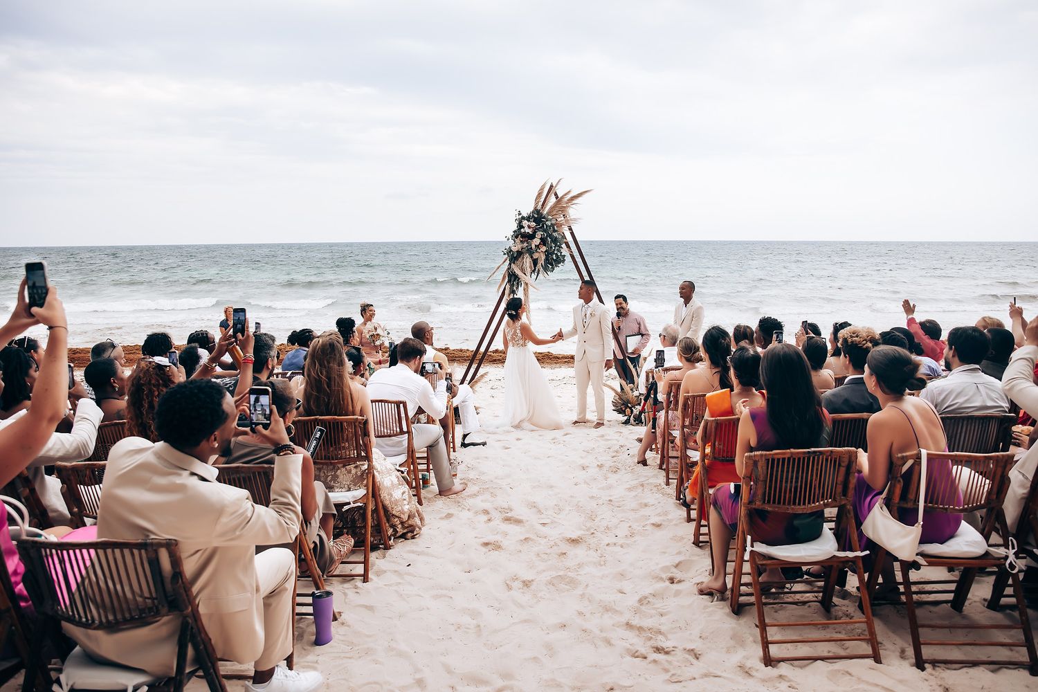 Guests seated in white chairs on a sandy beach for an oceanfront wedding ceremony.