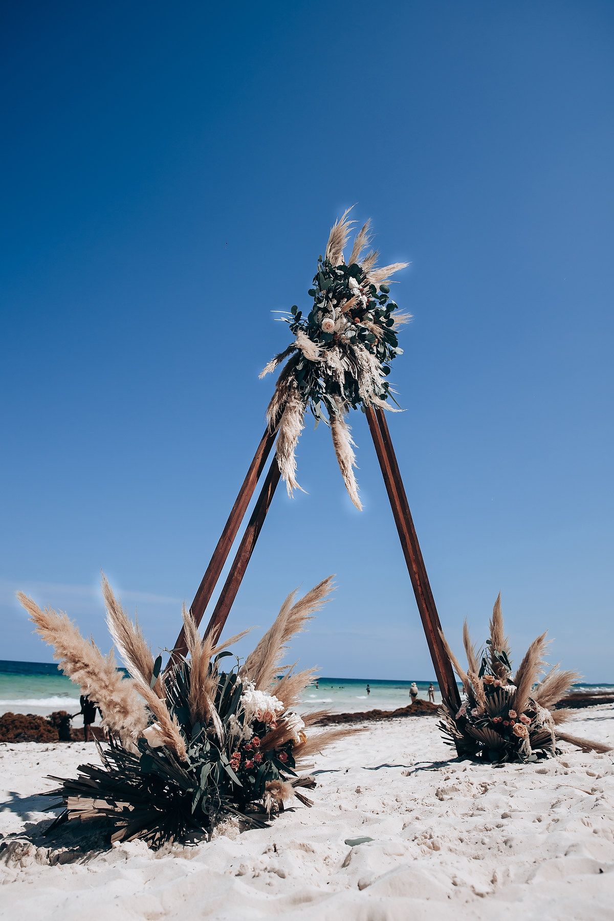 Rustic wooden teepee arch decorated with pampas grass and florals stands elegantly on a white sandy beach.