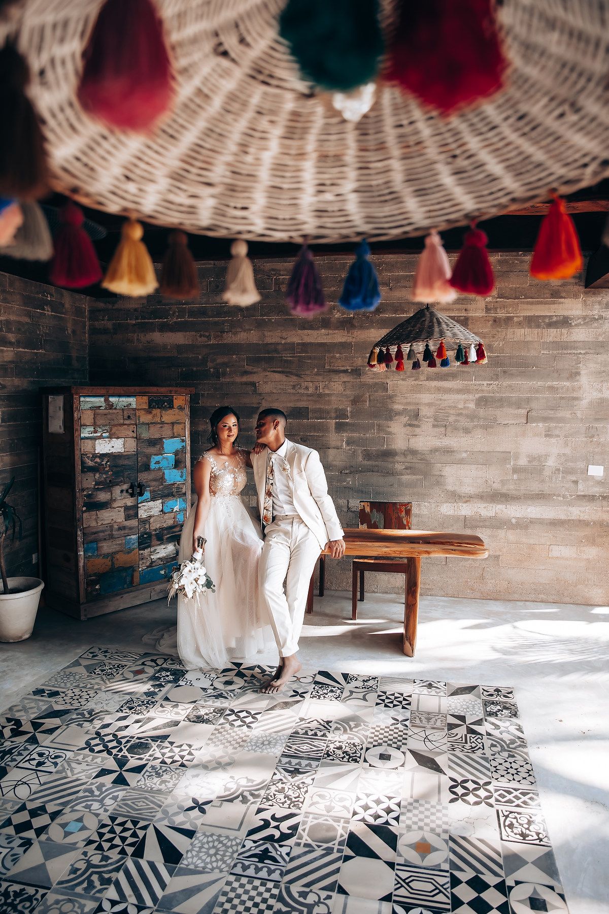 Wedding couple poses on patterned floor tiles under decorative hanging basket with colorful tassels.