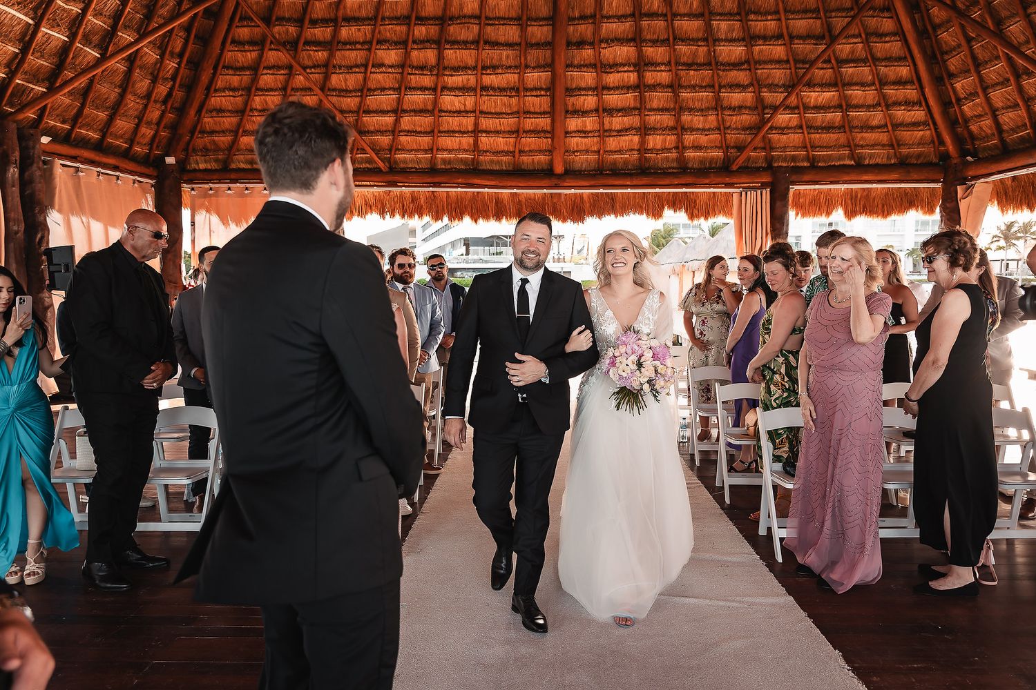 Wedding reception guests dancing under a rustic thatched roof pavilion.