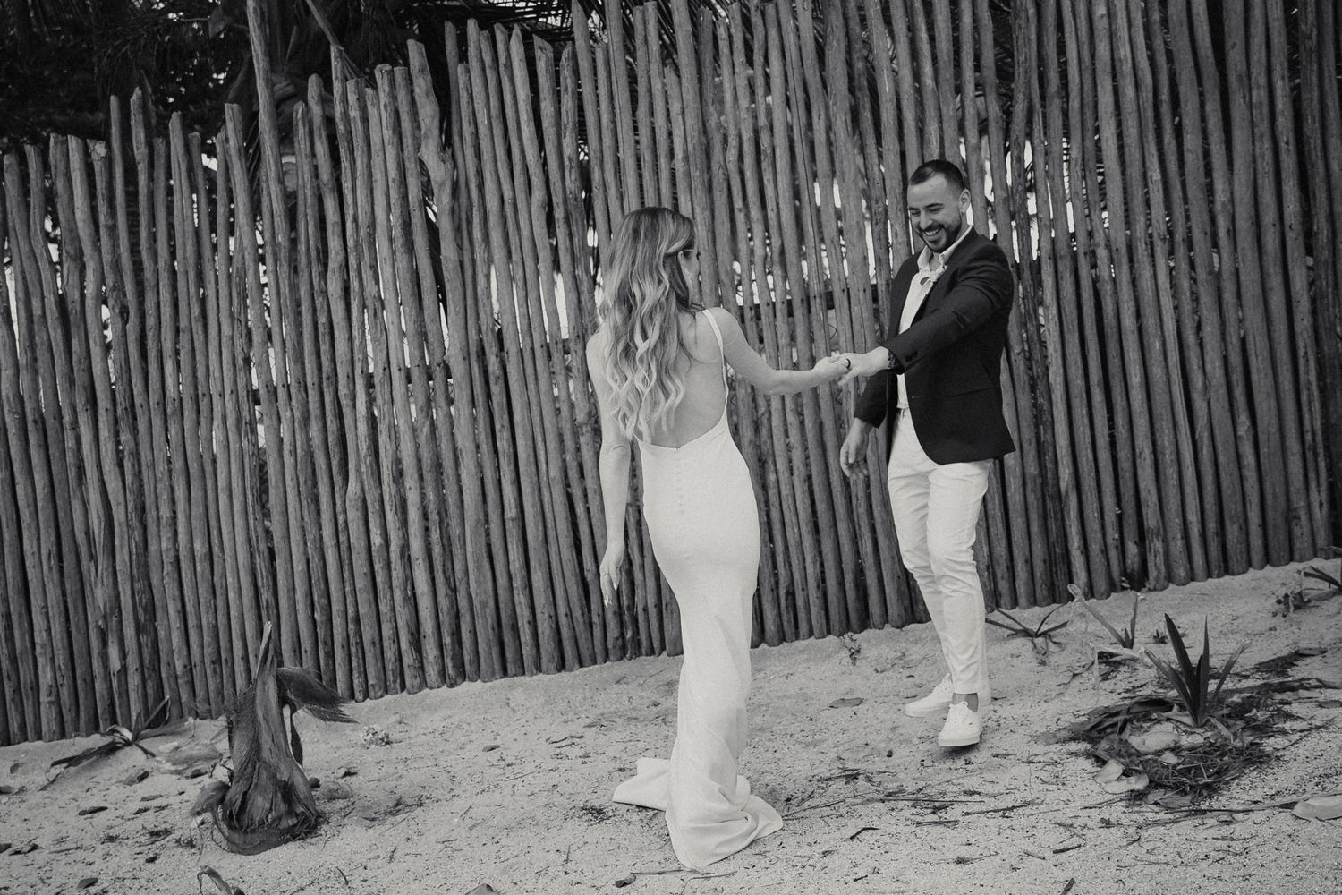Bride and groom share a romantic moment by a rustic wooden fence on sandy beach in black and white.
