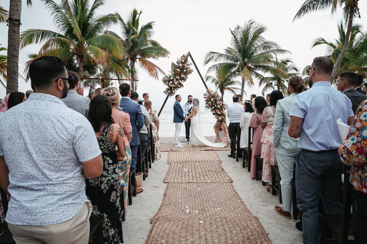 Couple exchanges vows at beach wedding ceremony with palm trees and triangular floral arch backdrop.