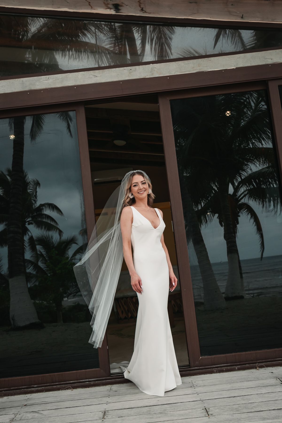 Bride in a sleek white wedding gown and veil stands smiling before large glass doors reflecting tropical palm trees.