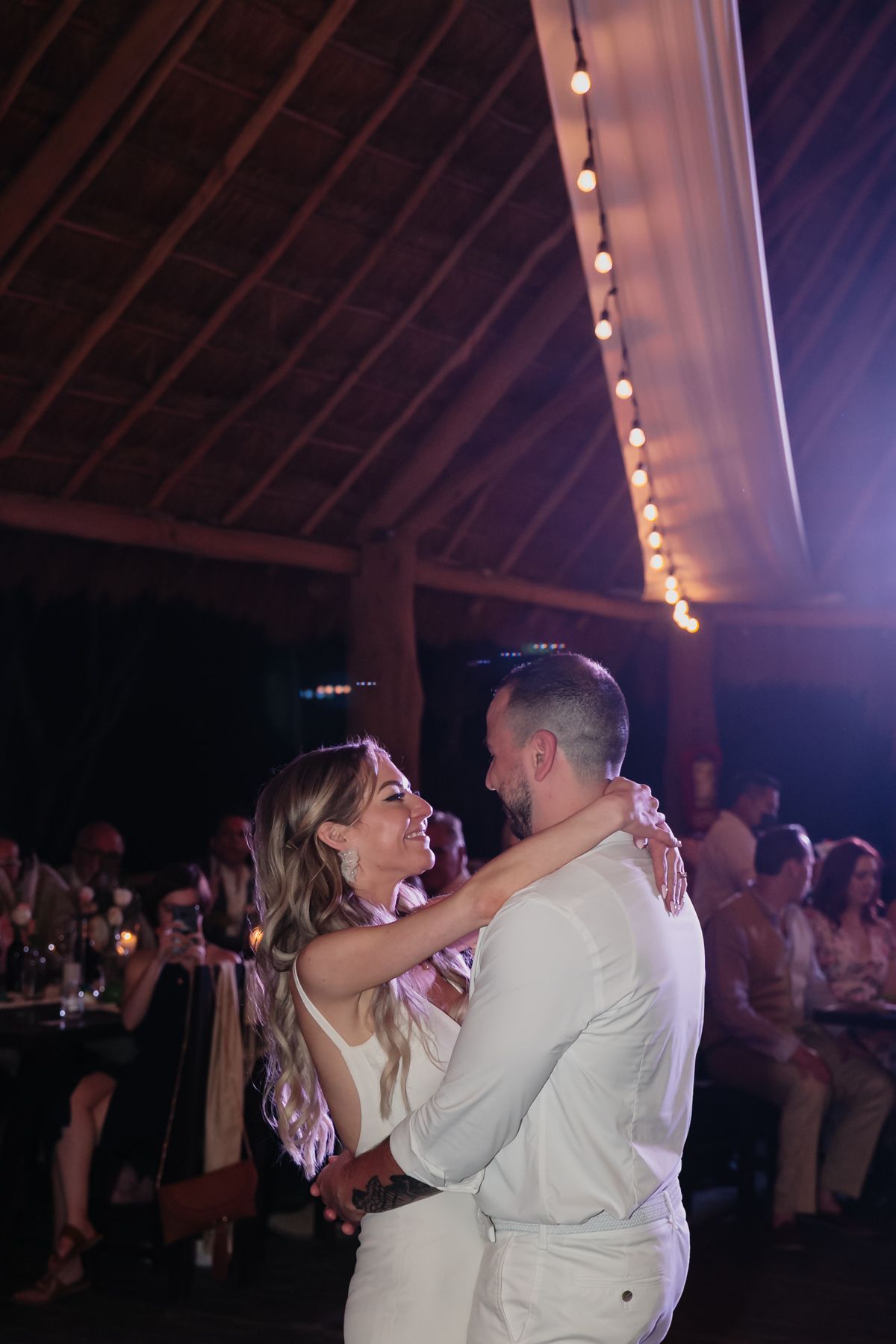 Bride and groom share romantic first dance under warm string lights in open-air wedding venue.