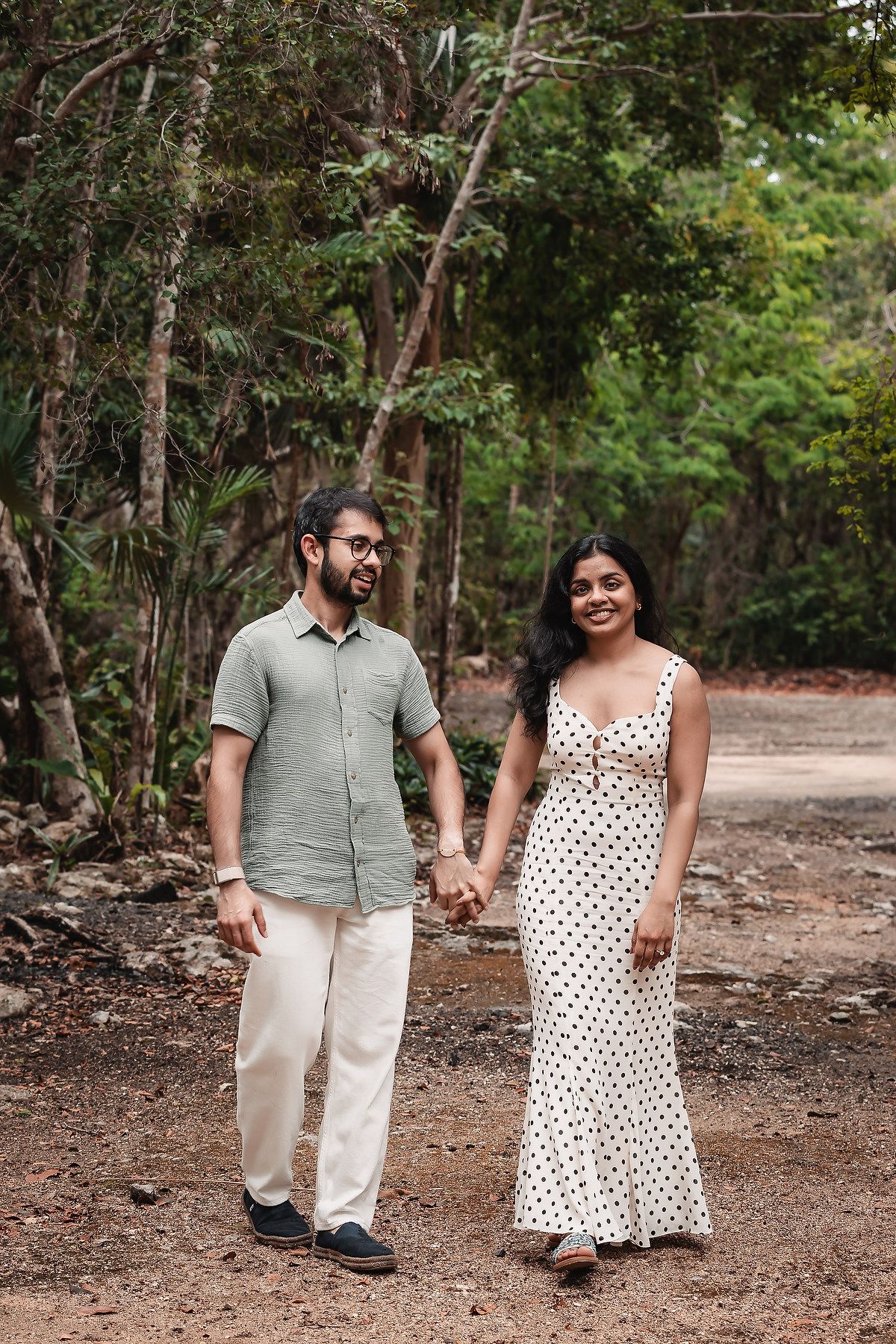 A couple in polka dot dress and sage shirt walk together on a forest trail.