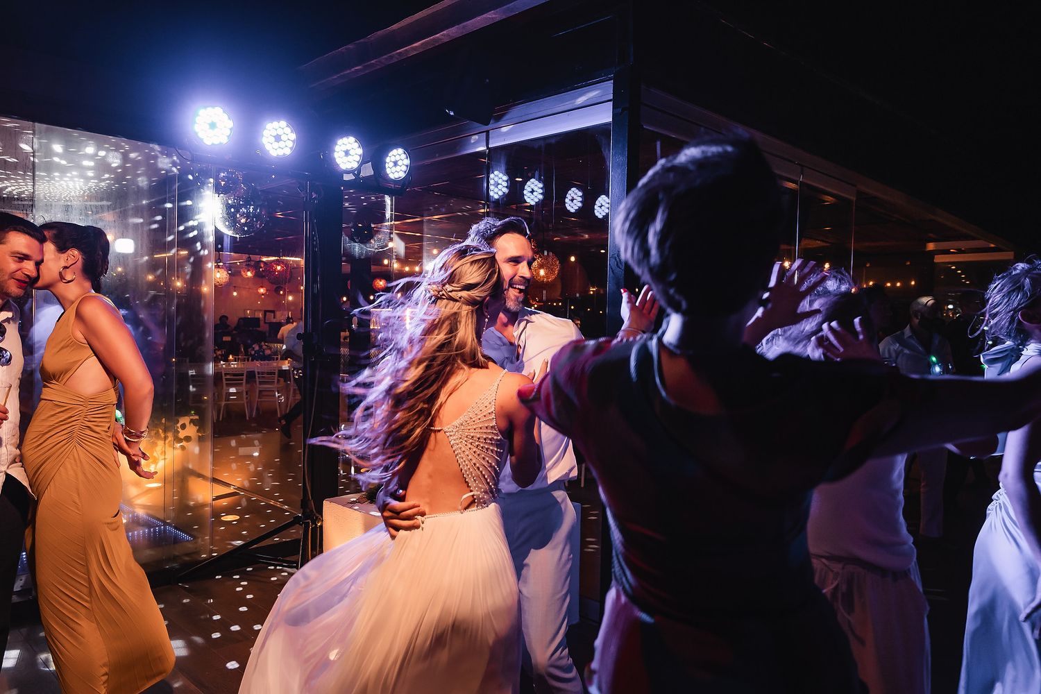 Wedding guests dance energetically on a dark dance floor illuminated by bright spotlights.