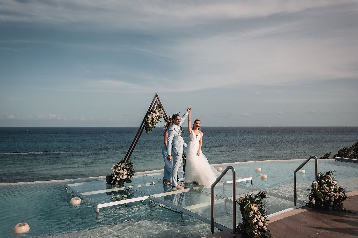 Couple celebrating under geometric floral arch on oceanfront deck during sunset ceremony.