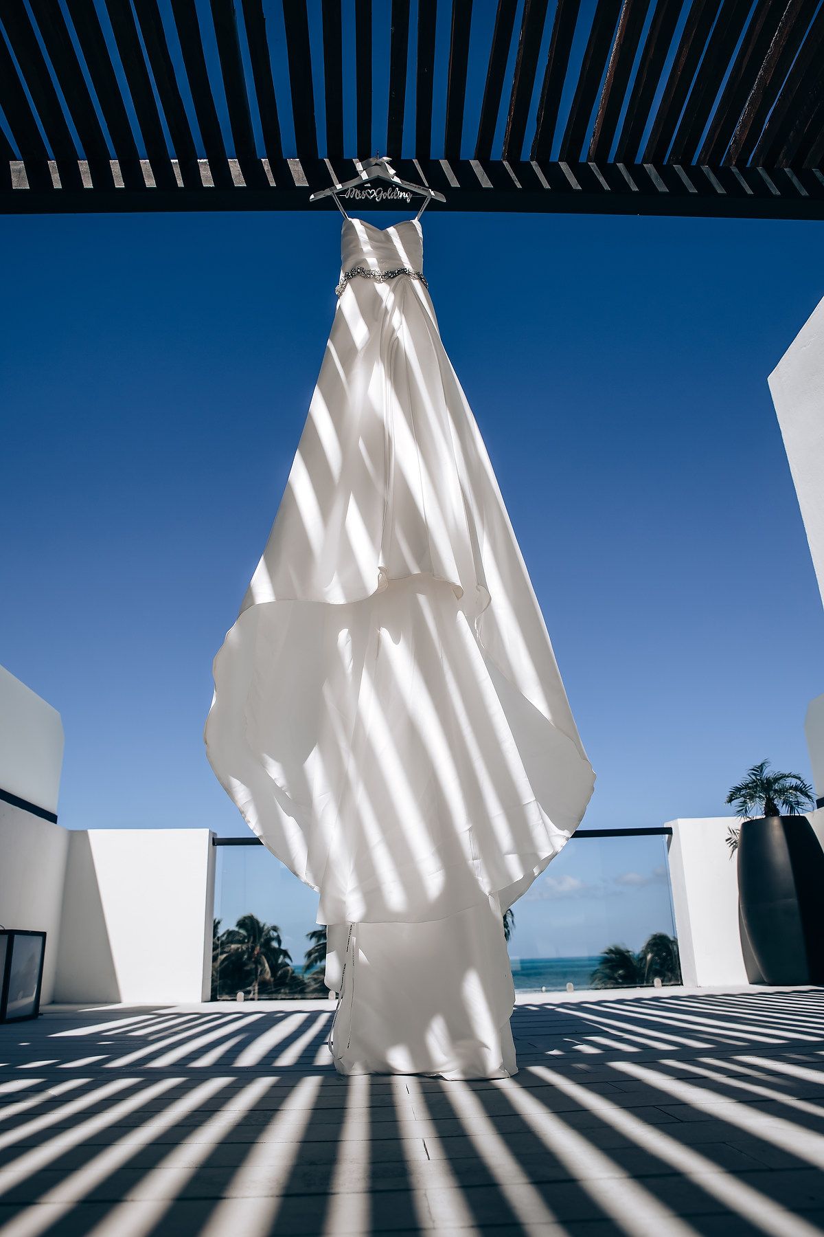 White wedding dress hanging elegantly against blue sky with dramatic shadows from pergola overhead.