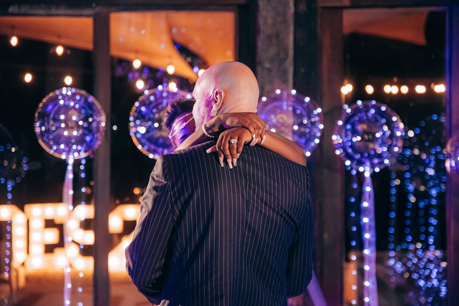 A couple embraces on a dance floor with glowing purple LED balloons and string lights creating a romantic atmosphere.