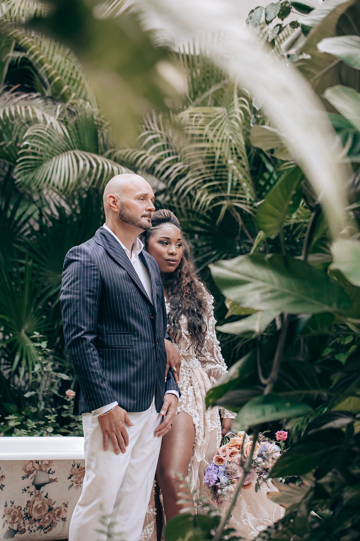 A couple poses elegantly against lush tropical foliage in formal attire during a romantic photoshoot.
