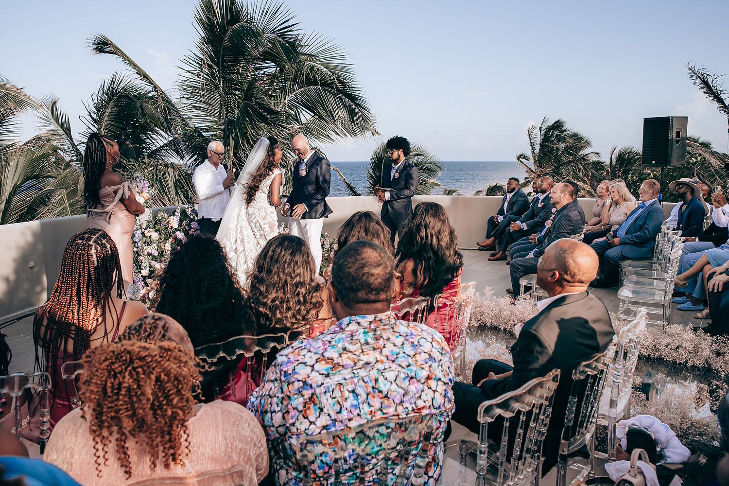 A group of people gather on a beach with palm trees and ocean views during a tropical celebration event.