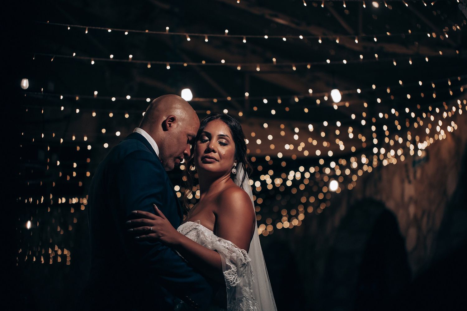 A romantic wedding couple dances beneath twinkling string lights at an outdoor evening reception.