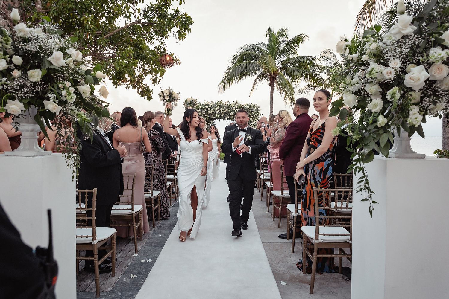 Wedding guests walk down a decorated outdoor aisle lined with white floral arrangements and greenery.