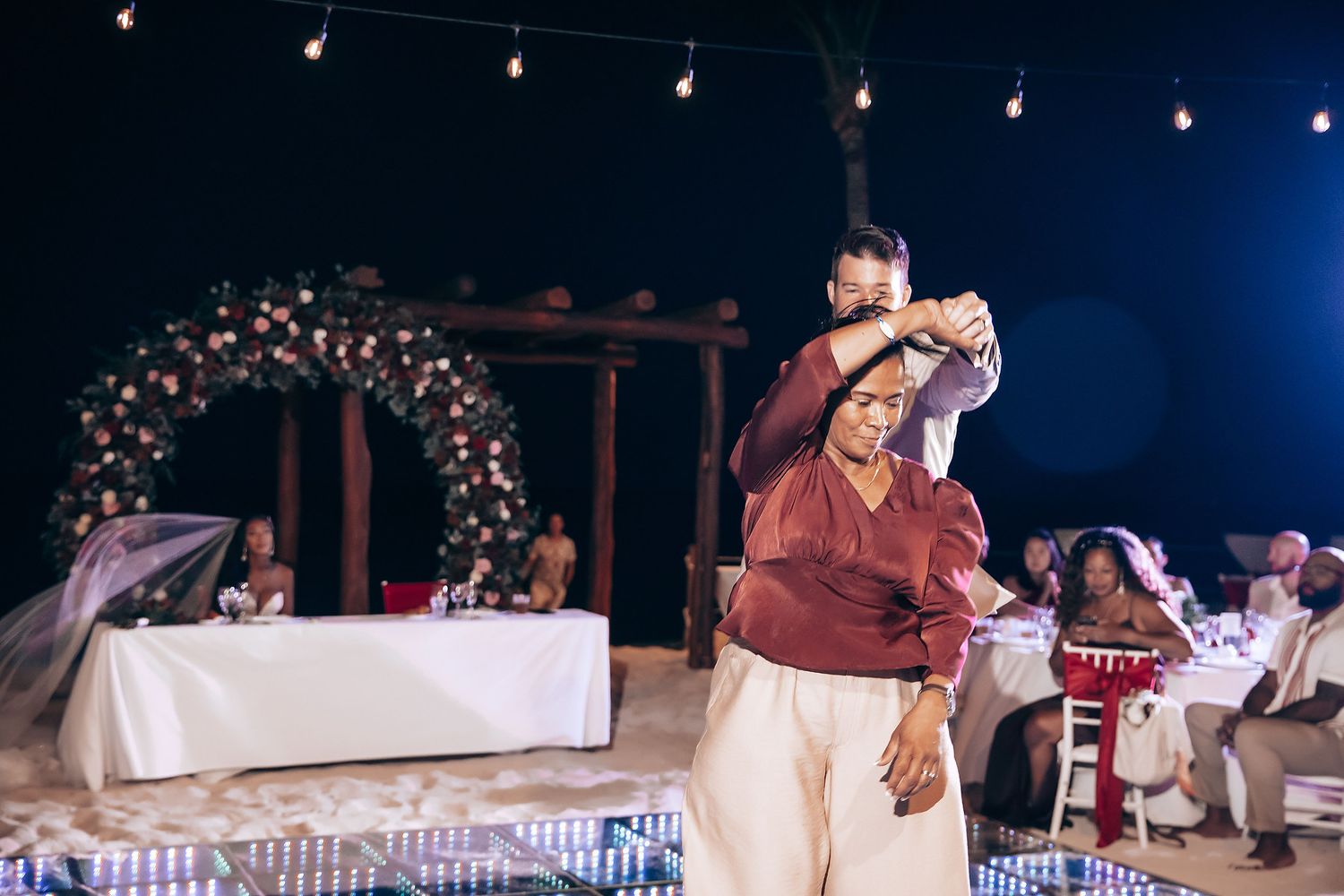 Wedding guests dance and mingle under string lights at an outdoor evening reception.