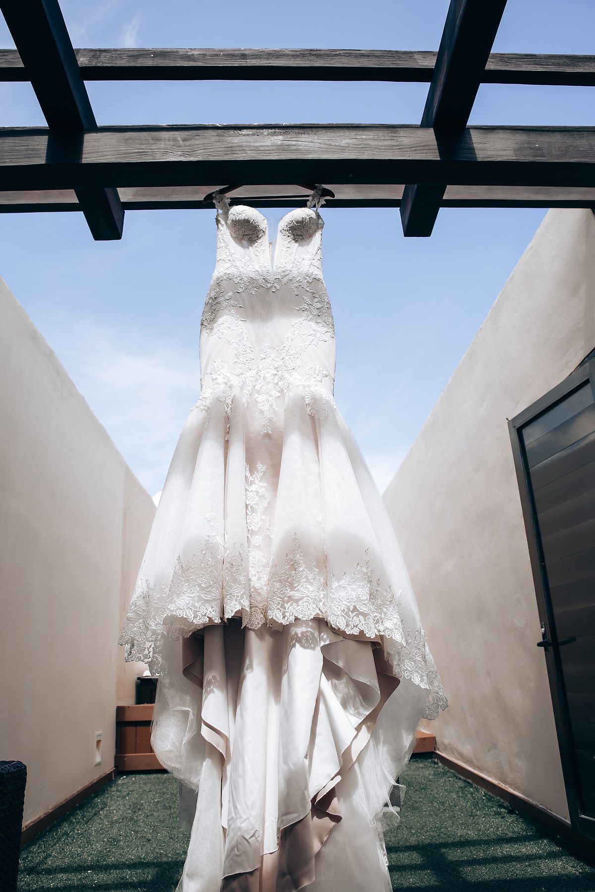 A white lace wedding dress with intricate details hanging from wooden beams against a bright sky.