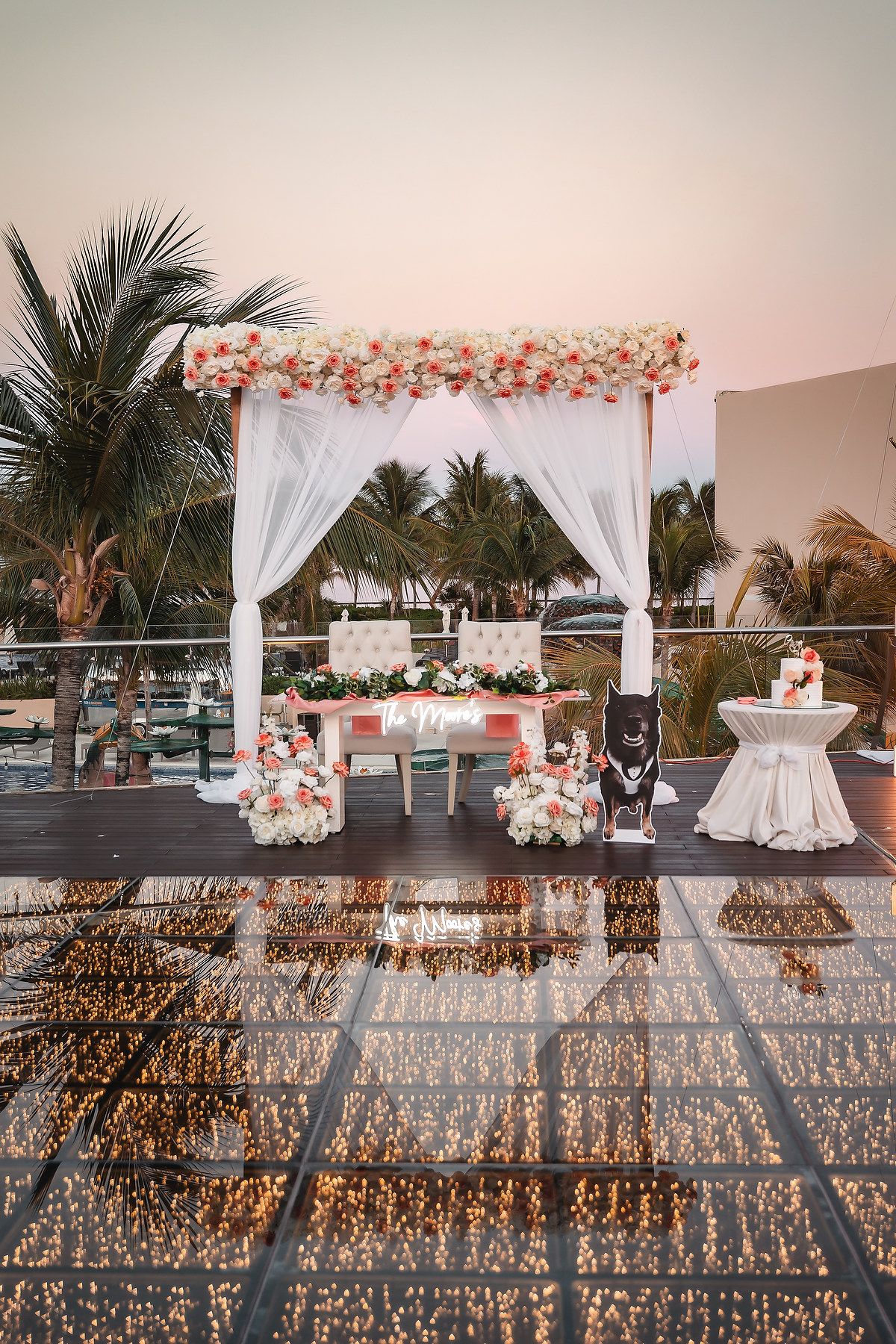 Wedding reception setup with white draped arch and coral flowers on a tropical terrace.