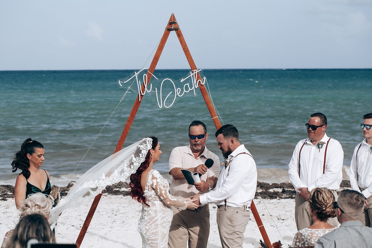 A beachside wedding ceremony takes place under a wooden triangular arch with turquoise ocean waters in the background.