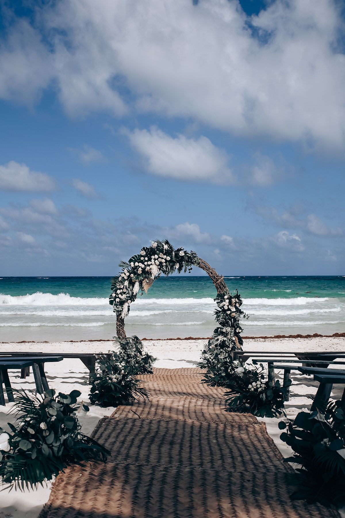 Wedding arch decorated with white flowers stands on a beach against turquoise waters and blue sky.