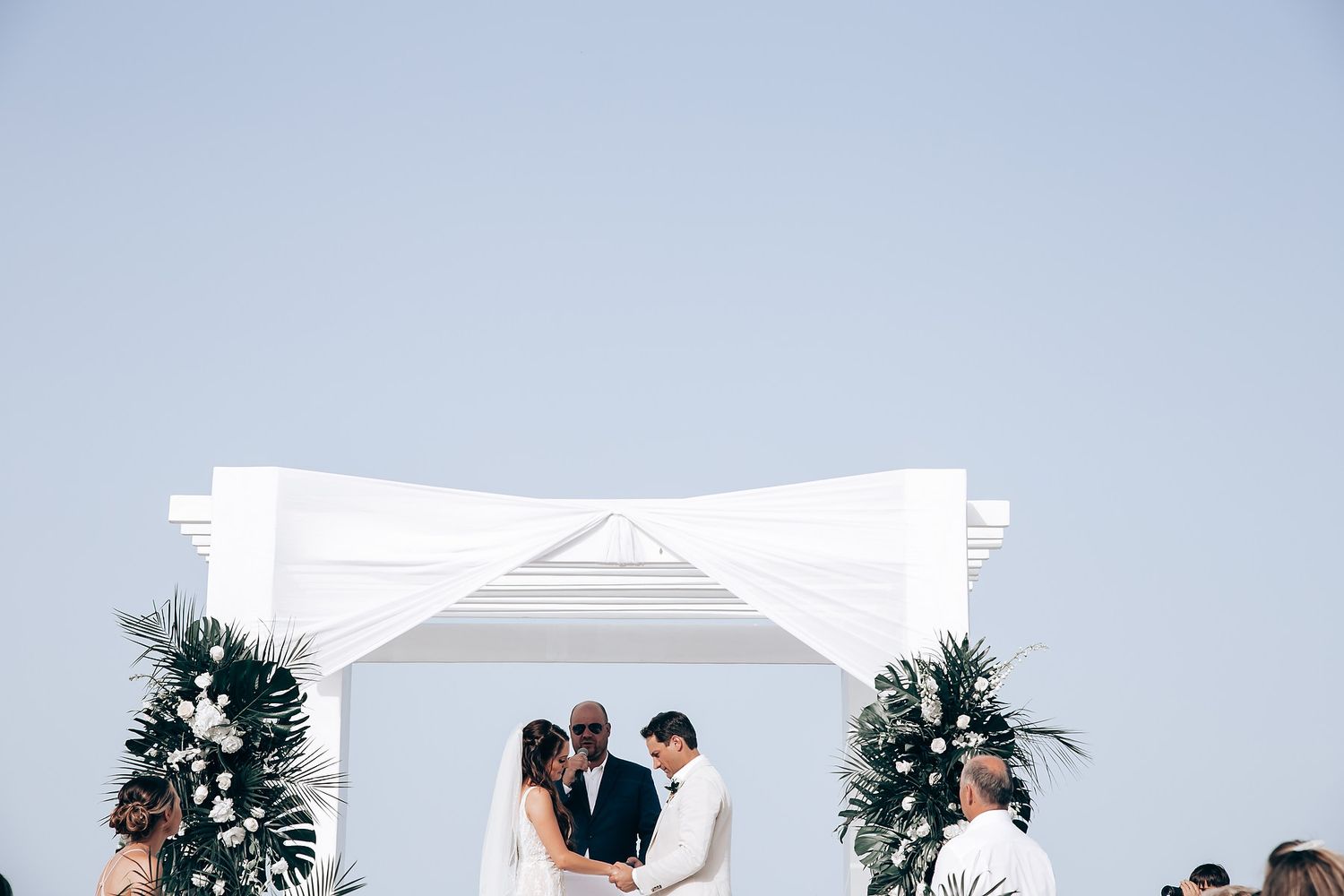 A romantic beach wedding ceremony setup with white chairs and palm frond decorations against an ocean backdrop.