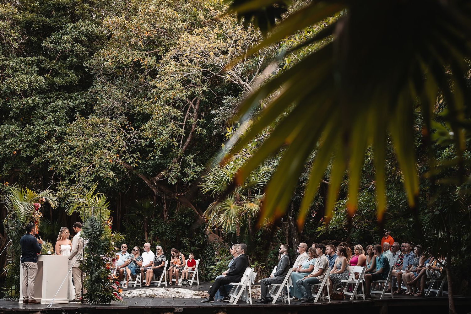 Outdoor wedding ceremony setup with white chairs surrounded by lush tropical foliage and palm fronds.