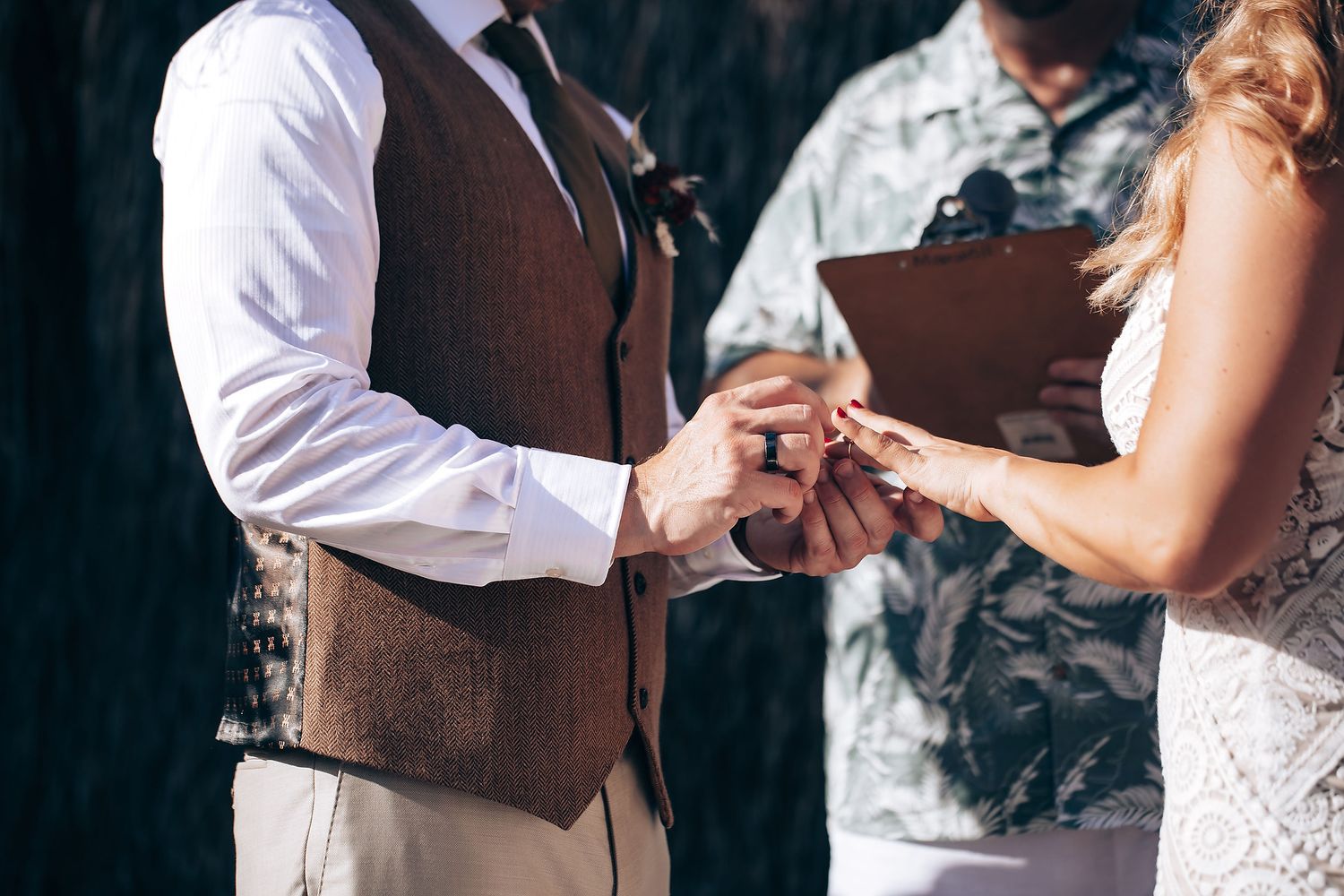 A close-up of hands exchanging wedding rings during a ceremony with a brown vest and white dress visible.