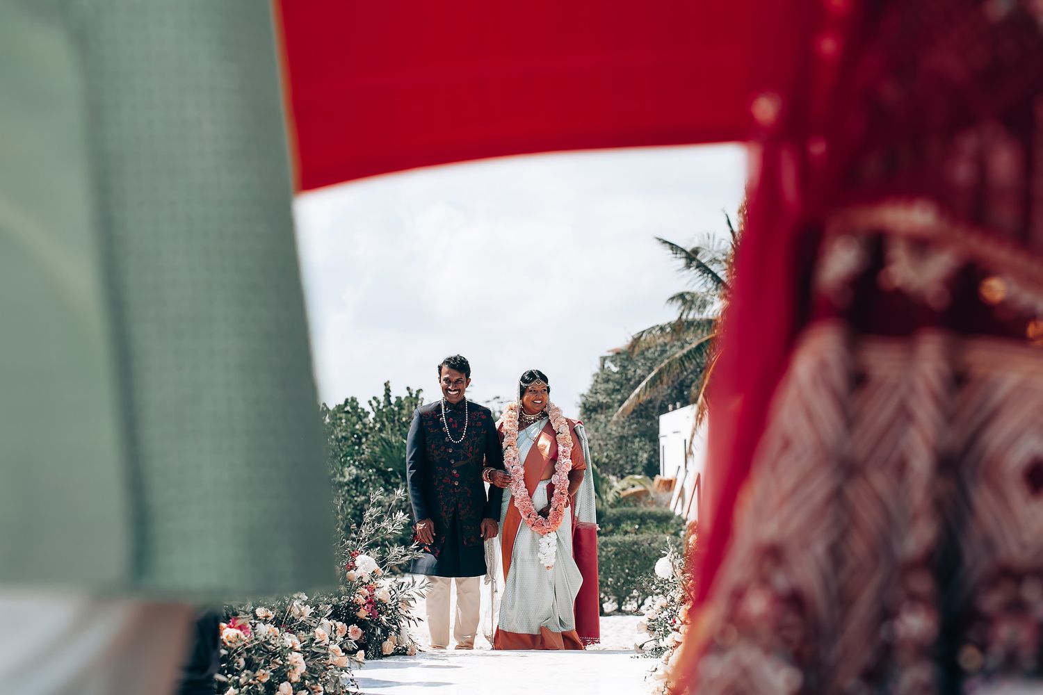 Partial view of wedding attendees in traditional red and mint green attire during a beach ceremony.
