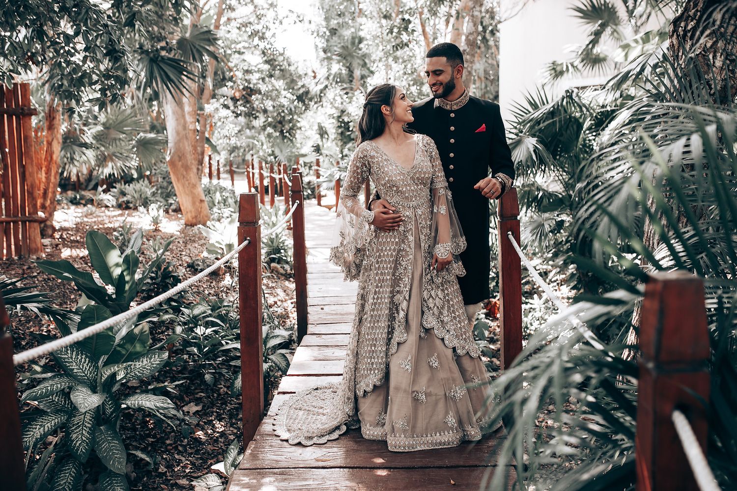 Wedding couple posing on elevated wooden walkway amidst tropical garden landscape.