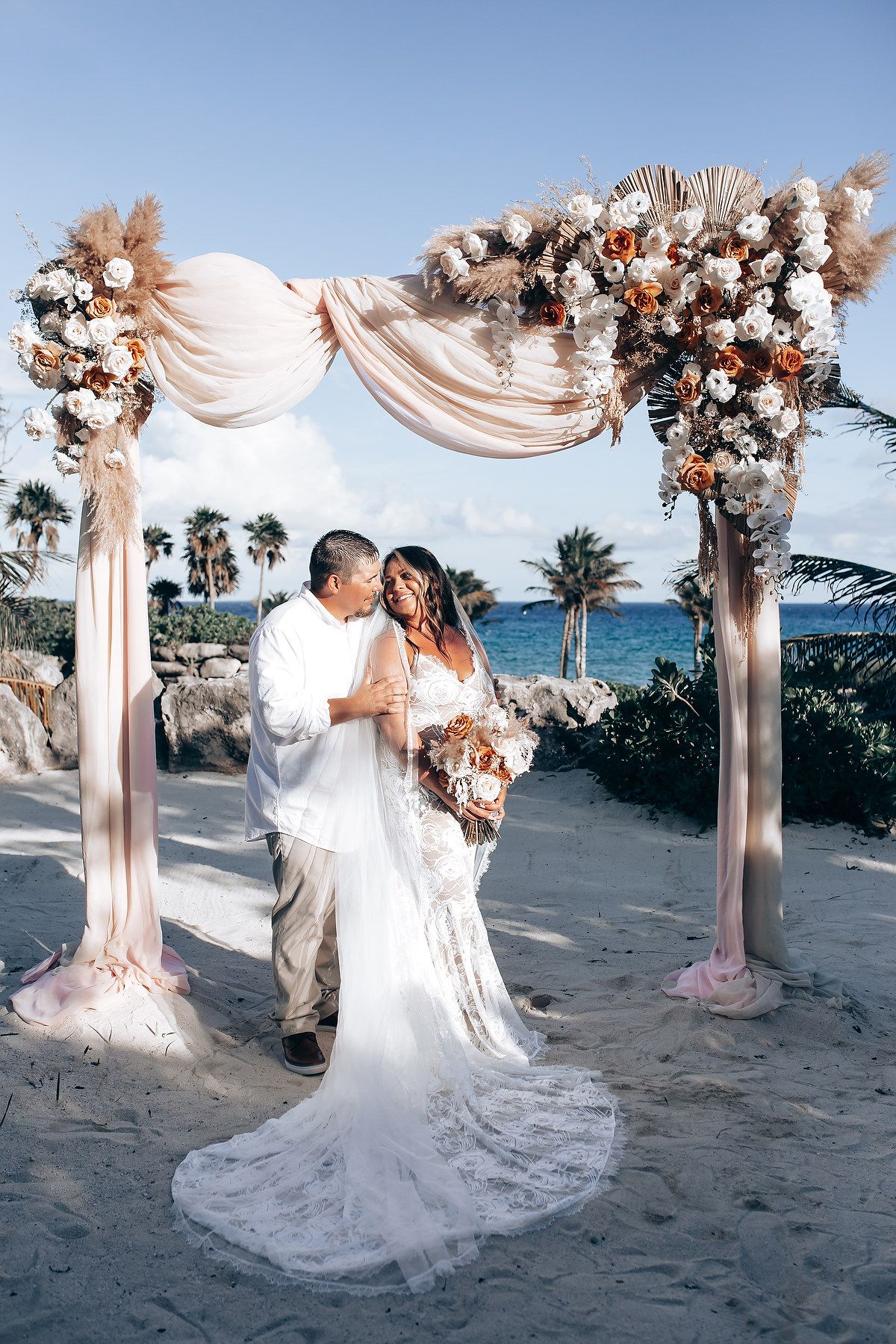 Romantic beach wedding ceremony setup with flowing fabric arch and floral decorations against ocean backdrop.