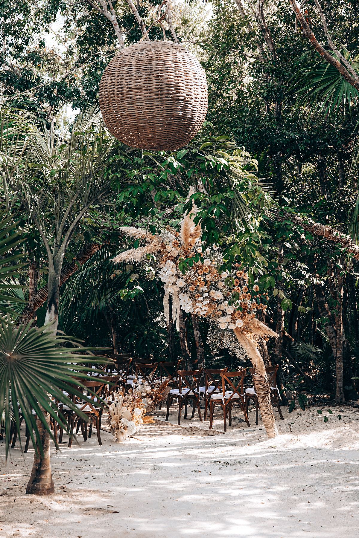 Rustic outdoor wedding venue with woven basket lanterns and tropical foliage in a beachside forest setting.