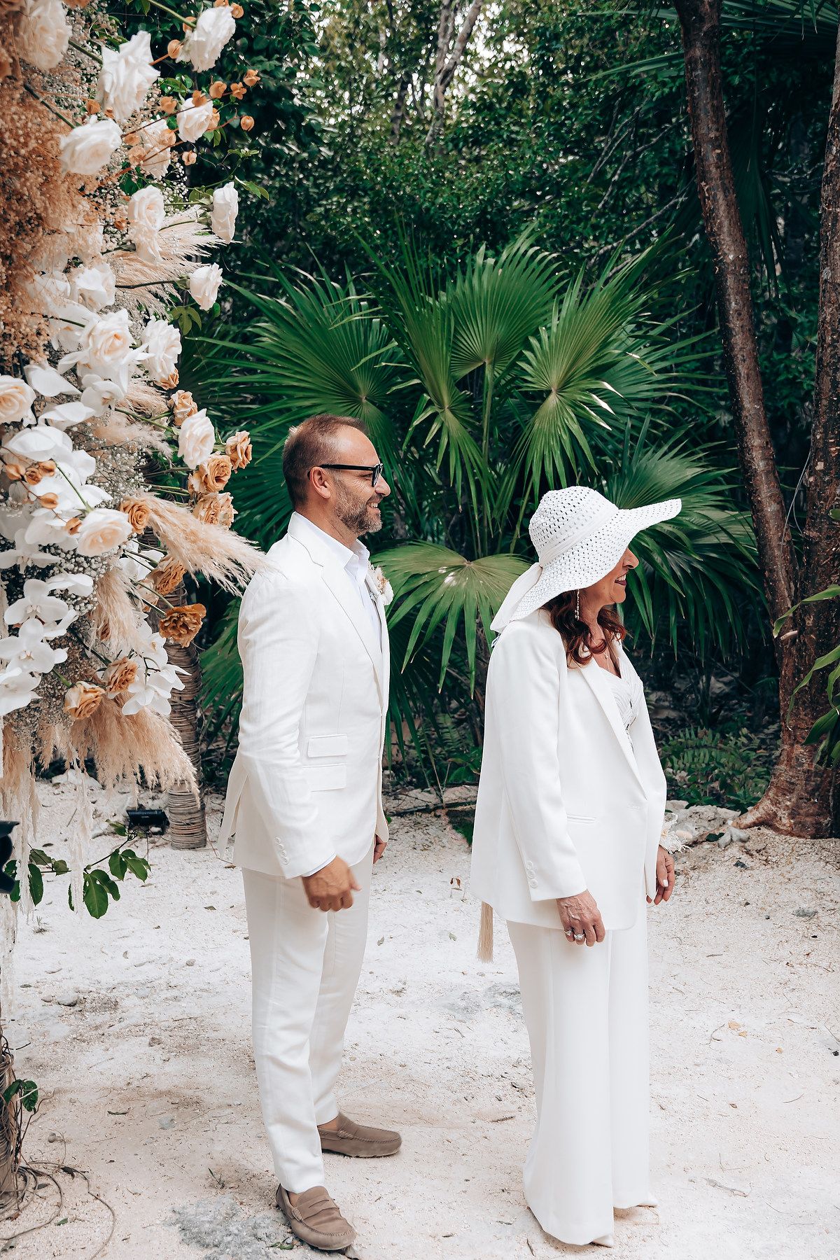 A couple in white attire walks along a sandy tropical path bordered by palm trees and lush greenery.