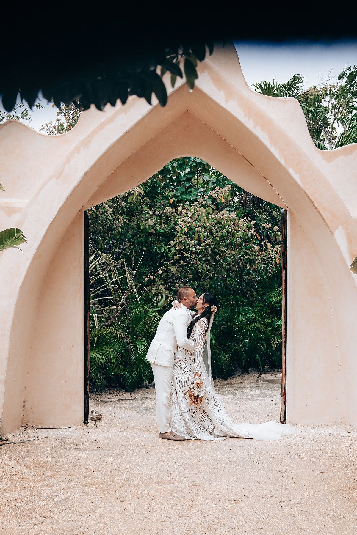 A romantic couple in white attire shares an intimate moment under a series of beige arched doorways on a sandy beach.