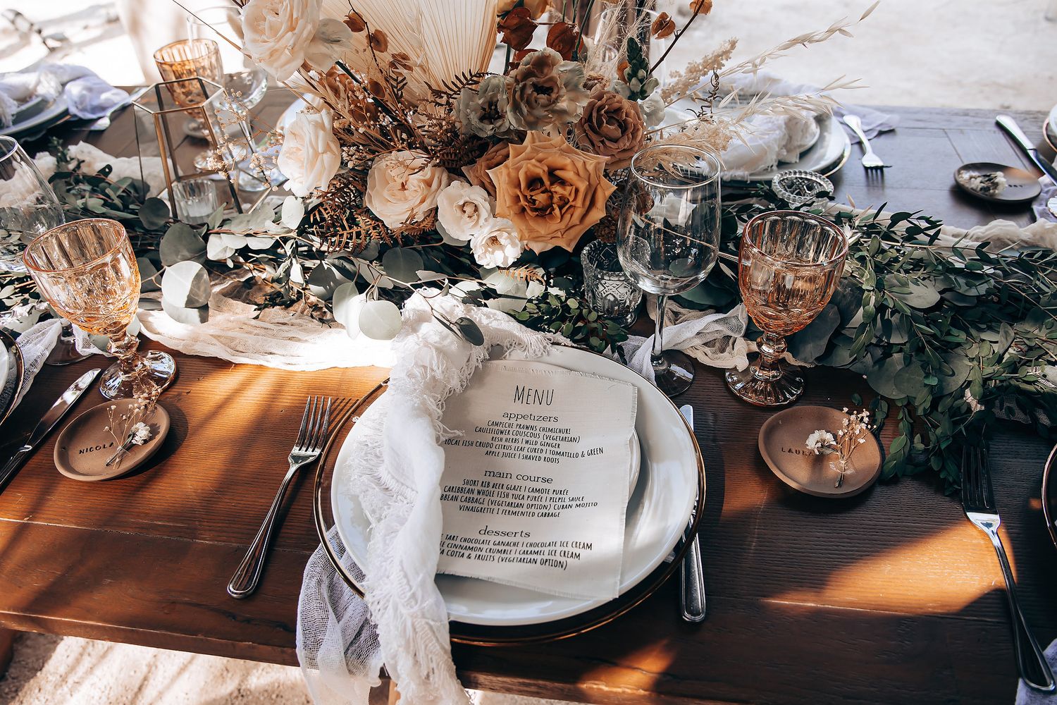 Close-up of wedding table settings with white plates, menus, and peach-colored floral arrangements on wooden tables.