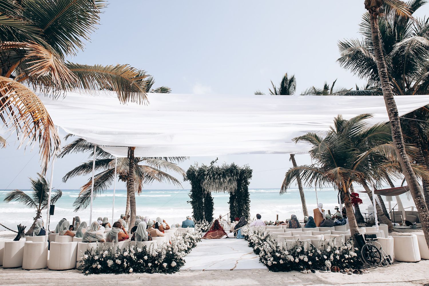 Tropical beach wedding venue with white chairs and floral arrangements under palm trees.