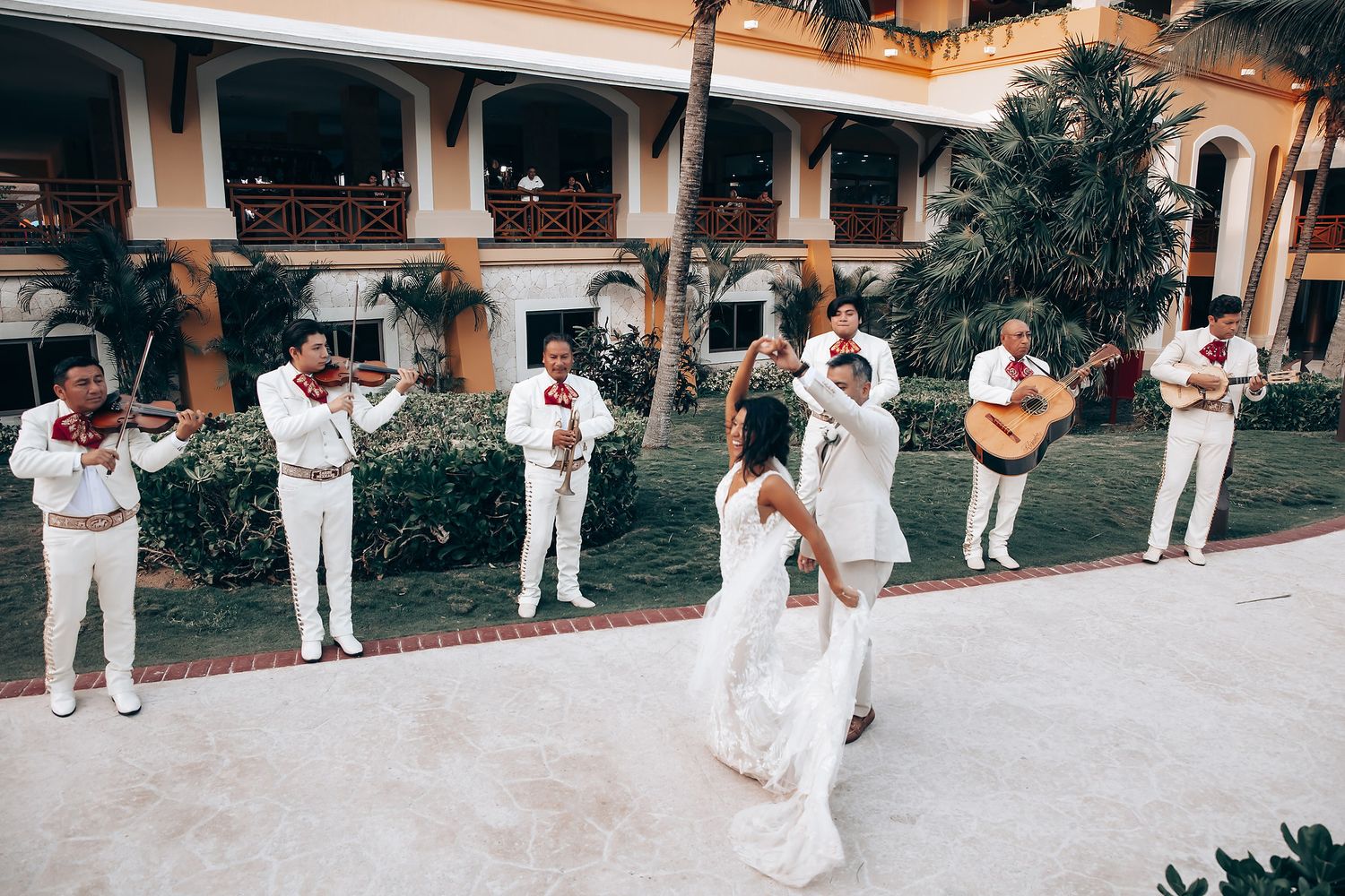 Wedding party members in white outfits dance and celebrate along a Spanish colonial style veranda.