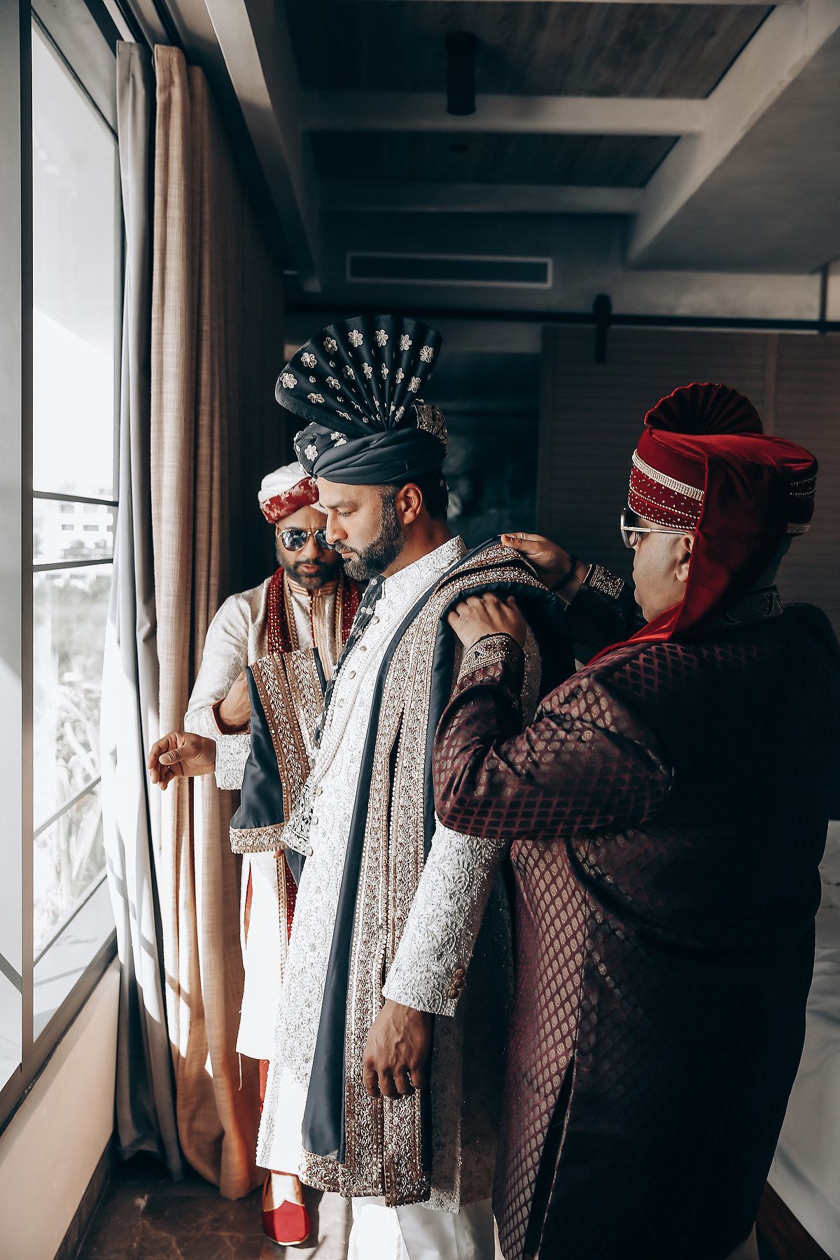 People in traditional Indonesian wedding attire stand together in an indoor setting with natural window light.