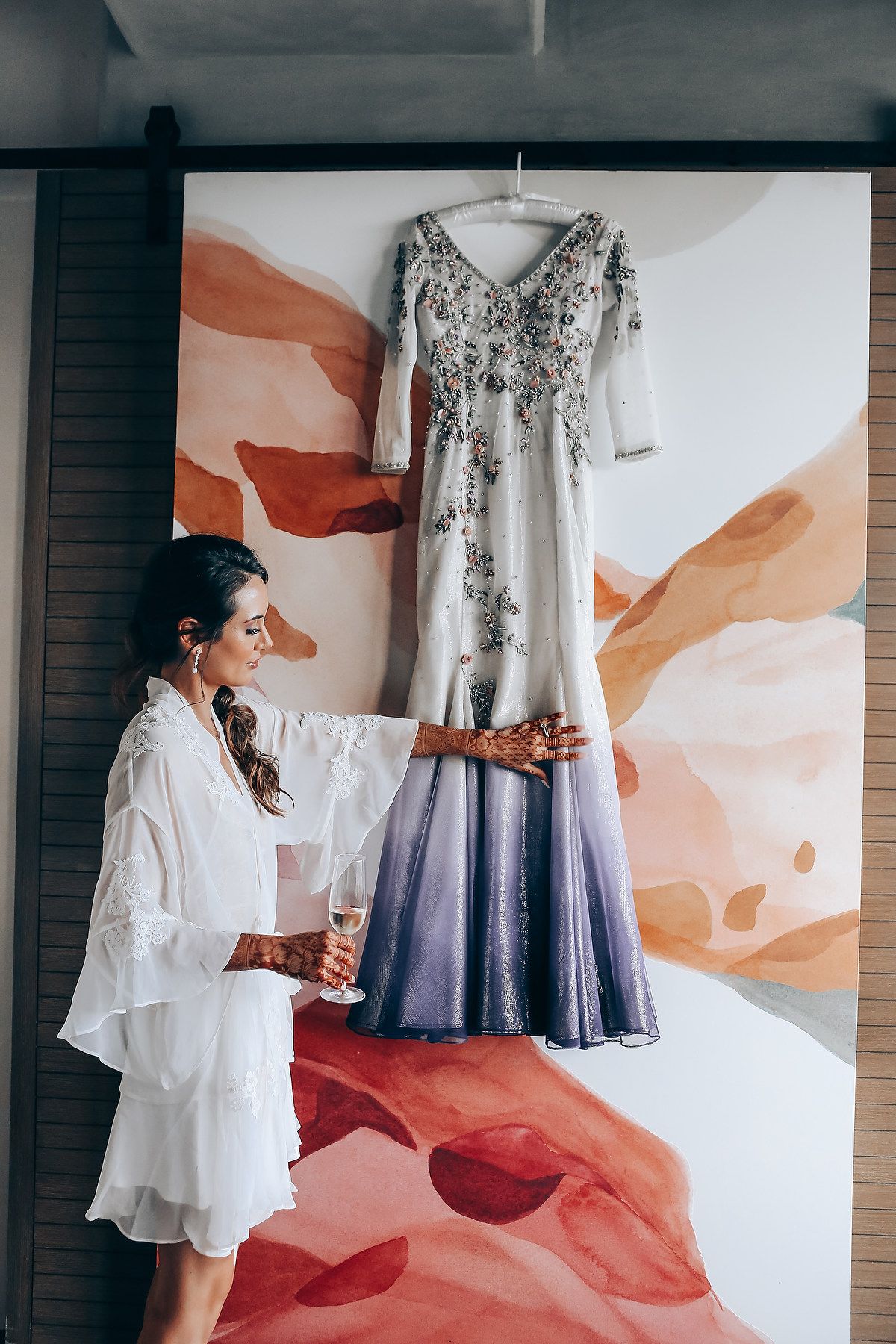 A flowing white and blue ombre wedding dress with floral embellishments hangs against a peach-colored abstract wall mural.