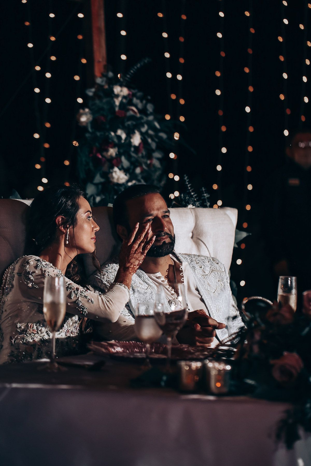 Two people in white lace outfits lounge on pillows while watching something in a dimly lit room with twinkle lights.