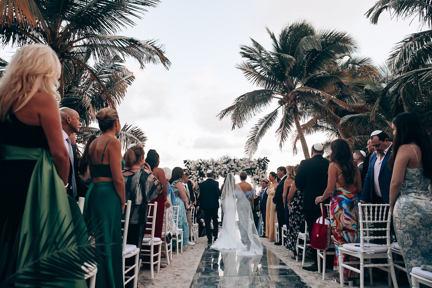 Wedding guests gathered on a beachfront venue with white chairs and tropical palm trees at dusk.