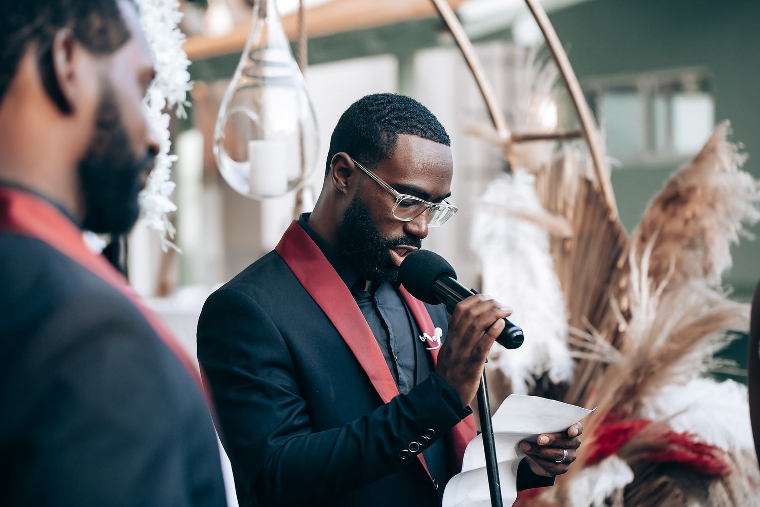 A person in a black suit with red lapels adjusts their tie while standing near decorative dried pampas grass arrangements.