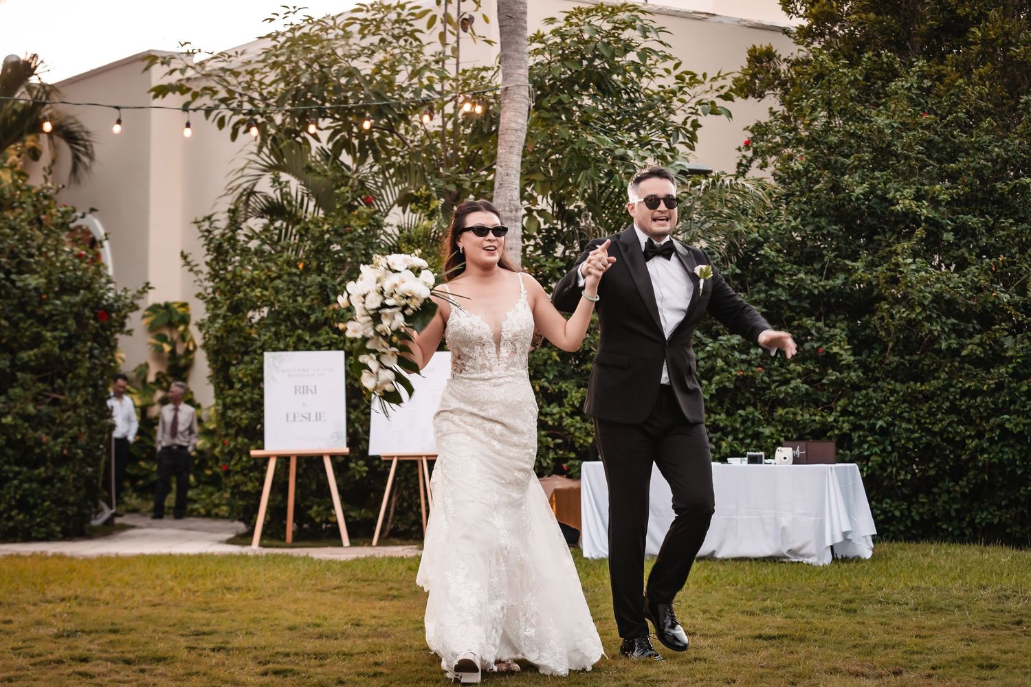 Wedding couple walks together on a garden path surrounded by lush greenery and decorative lighting.